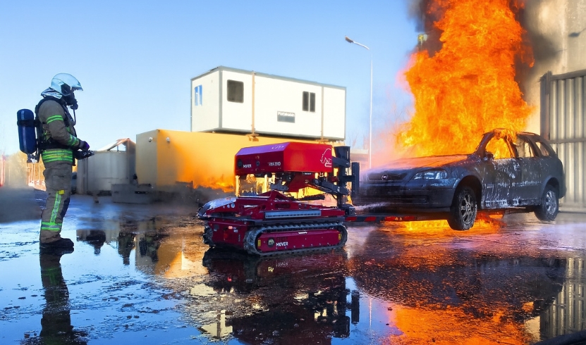 Remote-controlled recovery robot towing burning car as firefighter supervises.
